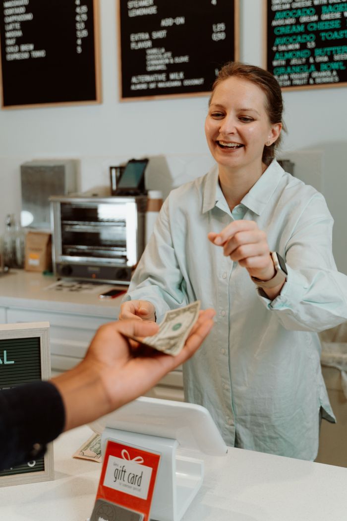 services-03 A cheerful barista receiving cash from a customer at a modern cafe counter, promoting a welcoming atmosphere.