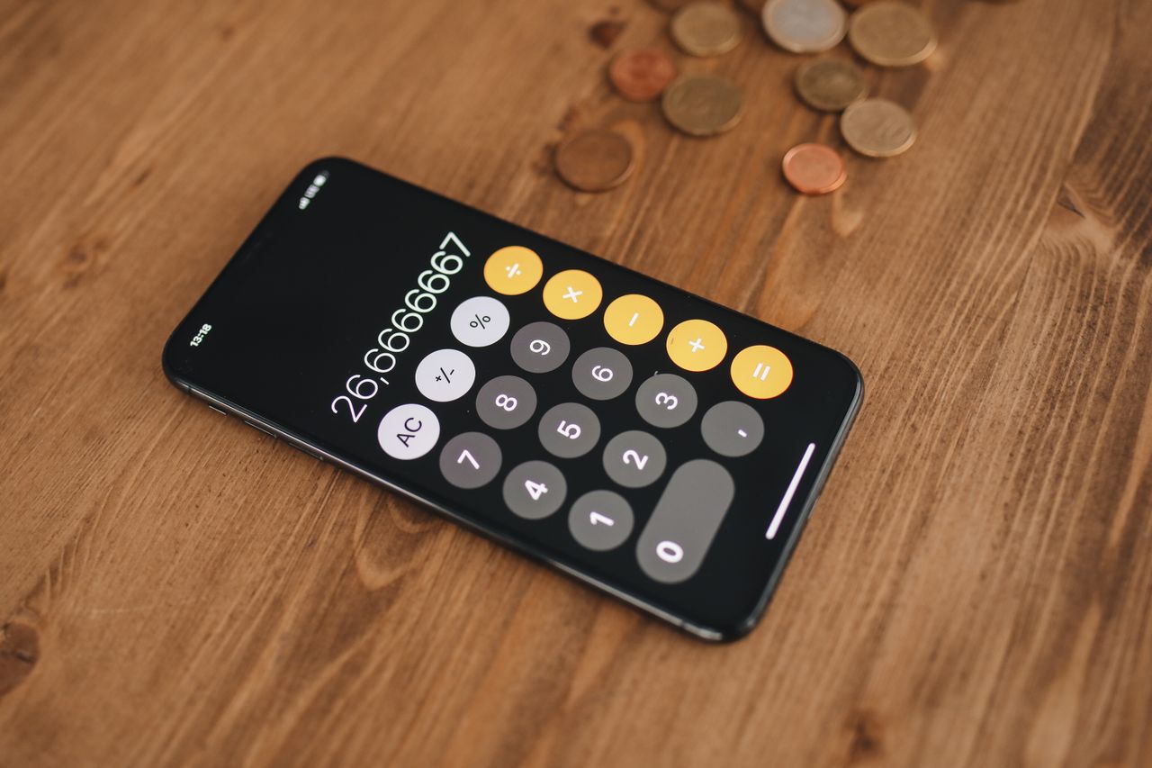 A close-up of a smartphone displaying a calculator app surrounded by coins on a wooden table.